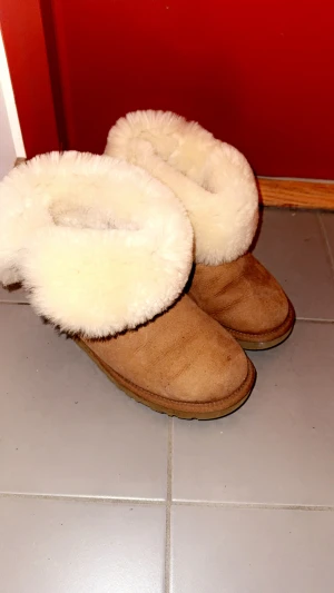 UGG boots med päls och knapp - Säljer ett par klassiska UGG boots i beige mocka med fluffig vit päls runt skaftet. Skorna har rund tå, platt sula och en snygg knappdetalj på sidan. Perfekta för kalla dagar och riktigt mysiga att ha på sig.