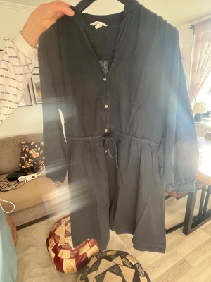 Mörkblå kort klänning med knyt - Snygg mörkblå kort klänning med knappar framtill och knyt i midjan. Klänningen har långa ärmar med lätt puff och är tillverkad i ett tunt, luftigt tyg som känns skönt mot huden. Perfekt för dig som gillar en avslappnad men ändå stilren look.