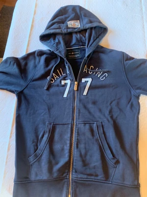 Marinblå hoodie från Sail Racing - Snygg marinblå hoodie från Sail Racing med dragkedja och huva. Framsidan har broderade vita detaljer med texten 'Sail Racing' och siffran 77. Tröjan har snörning i huvan och två stora fickor framtill. Perfekt för chill dagar och sportig stil.              OBS på vänstra arm finns det ett slags skada. 