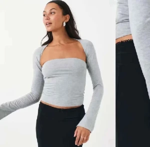 Grå croppad bolerotopp med långa ärmar - Säljer en ljusgrå croppad bolerotopp med långa ärmar och axelbandslös design. Toppen har en stilren och modern look med smal passform som framhäver siluetten. Perfekt för dig som gillar minimalistisk stil och vill ha något trendigt.