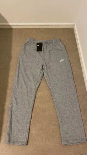 Grå mjukisbyxor från Nike - Grå mjukisbyxor från Nike med elastisk midja och klassisk logga på vänster lår. Byxorna har en bakficka med knapp och är tillverkade i 80% bomull och 20% polyester för skön komfort. Perfekta för chill eller träning.