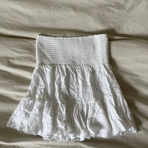 Vit volangkjol med smock - Supersöt vit kortkjol med smock i midjan och volang nertill. Kjolen har en luftig och lätt känsla, perfekt för varma dagar. Passar dig som gillar en feminin och trendig stil.