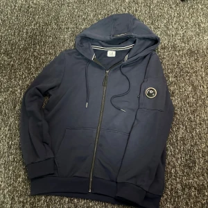 Marinblå hoodie från C.P. Company - Säljer en marinblå hoodie från C.P. Company med dragkedja, huva och snörning. Tröjan har en klassisk patch med logga på ärmen och stora fickor framtill. Perfekt för dig som gillar streetwear och vill ha en stilren look.