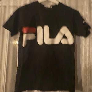 Svart FILA t-shirt med stor logga - Svart t-shirt från FILA i storlek XS med stor vit och röd logga framtill. Klassisk rund halsringning och korta ärmar. Perfekt för dig som gillar sportiga och stilrena plagg med tydlig branding.