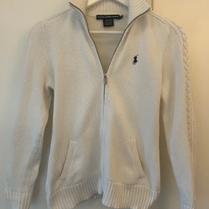 Vit stickad kofta från Ralph Lauren - Snygg vit stickad kofta från Ralph Lauren Sport med dragkedja och ribbade muddar. Tröjan har långärmade ärmar, två fickor fram och ett mörkblått broderat Polo-logo på bröstet. Flätad detalj längs axlarna och tillverkad i 100% bomull. Köpt på plick för 1099kr, säljer då tröjan är för liten på mig. Storlek S men sitter som XS.