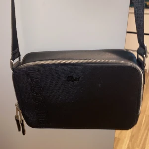 Svart axelväska från Lacoste - Snygg svart axelväska från Lacoste med präglad logga på framsidan och silverfärgad dragkedja. Väskan har en stilren, rektangulär form och justerbar axelrem. Tillverkad i tåligt syntetmaterial och har ett rymligt huvudfack med innerficka för småsaker.