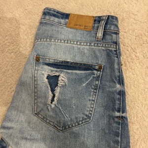 Blå slitna jeansshorts från Gina Tricot  - Säljer ett par blå jeansshorts från Gina Tricot med råa kanter och stora slitningar både fram och bak. Klassisk femficksmodell med dragkedja och knapp. Perfekt för en avslappnad och trendig sommarstil.
