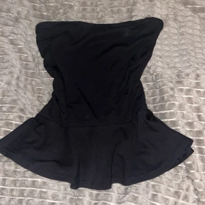 tubtopp med volang - Snygg svart tubtopp med volang nertill. Klänningen har en enkel och stilren design som ger en cool och trendig look. Perfekt för dig som gillar minimalistiska plagg med en feminin touch.