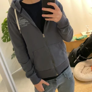 Mörkgrå hoodie från Superdry - Säljer en mörkgrå hoodie från Superdry med dragkedja, huva med vita snören och två fickor framtill. Klassisk logga på bröstet och mjukt material som passar perfekt till chill dagar. Snygg och enkel att matcha med jeans eller joggers.