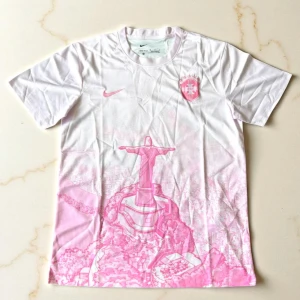 Vit och rosa Nike fotbollströja - Snygg vit fotbollströja från Nike med rosa detaljer och tryck av Kristusstatyn i Rio de Janeiro. Tröjan har klubbmärke på bröstet och klassisk rund hals. Tillverkad i lätt och ventilerande material, perfekt för träning eller match.