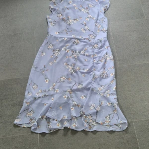 Ljusblå blommig klänning från NLY Trend - Superfin ljusblå klänning från NLY Trend med blommigt mönster i vitt och orange. Klänningen har en feminin passform, volangdetalj nertill och är ärmlös. Perfekt för sommarens alla tillfällen!