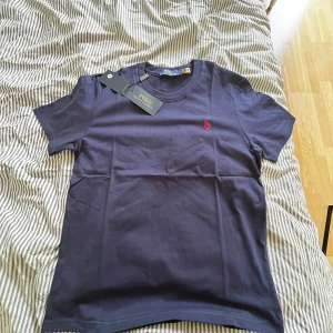 Mörkblå t-shirt från Polo Ralph Lauren - Snygg mörkblå t-shirt från Polo Ralph Lauren med klassisk röd logga broderad på bröstet. T-shirten är kortärmad och har en rund halsringning. Tillverkad i mjuk bomull för en skön känsla och stilren look.