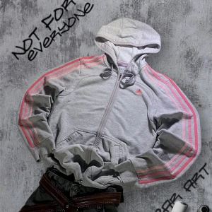 Retro Hoodie från Adidas rosa ärmar  - Säljer en grå huvtröja från Adidas med rosa ränder längs ärmarna. Tröjan har en dragkedja framtill och en justerbar huva med snören. Perfekt för en sportig look eller en avslappnad dag. Storlek M - Passar mer S/XS