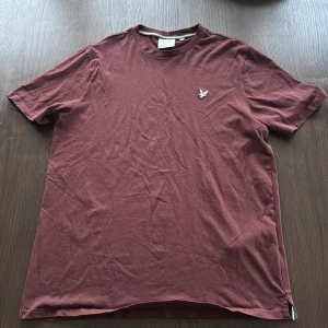Vinröd t-shirt från Lyle & Scott - Vinröd t-shirt från Lyle & Scott i bomull med klassisk passform. T-shirten har rund halsringning och en broderad grå logotyp med fågel på bröstet. Enkel och stilren design som funkar till det mesta.