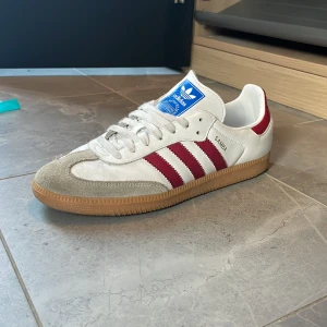 Adidas Samba vita med röda ränder - Adidas Samba sneakers i vitt skinn med klassiska röda ränder på sidorna och beige mockadetaljer vid tån. Skon har en brun gummisula och blå Adidas-logga på plösen. Perfekt för dig som gillar retrostil och streetwear.