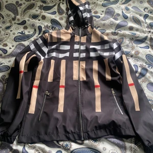 Burberry vindjacka med beige och svart mönster Ny med lappen kvar - Snygg vindjacka från Burberry med ikoniskt rutigt mönster i svart, beige, vitt och röda detaljer. Jackan har huva, dragkedja framtill och två fickor med dragkedja. Perfekt för dig som vill sticka ut med klassisk streetstyle.