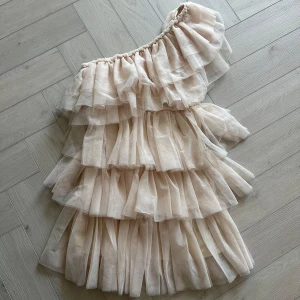 Beige oneshoulder klänning - Säljer en elegant beige oneshoulder klänning med flera lager av volanger. Perfekt för en stilfull kväll. Klänningen har en luftig och romantisk känsla.