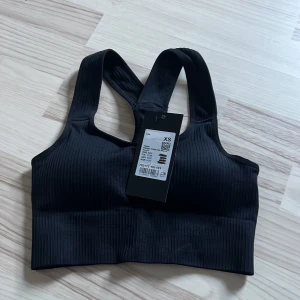 Svart ribbad sporttopp från TriDri. - Svart ribbad sporttopp från TriDri i sömlös design. Toppen har breda axelband och en tight passform som ger bra stöd vid träning. Materialet är stretchigt och bekvämt, perfekt för gymmet eller yoga. Enkel och stilren look med diskreta ribbade detaljer.