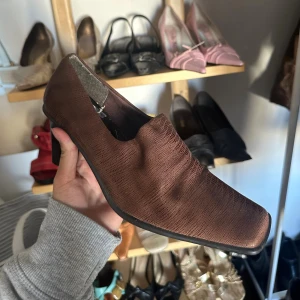 Bruna pumps med fyrkantig tå - Säljer ett par bruna pumps med fyrkantig tå och låg klack. Skorna har ett subtilt mönster som påminner om reptil och är tillverkade i ett material som ser ut som skinn. Perfekta för dig som gillar retrovibbar och vill sticka ut med en unik form.