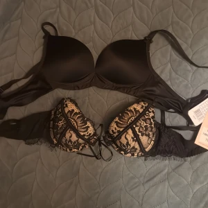 2 stycken Svart bh Freya & spets Forever Young - Två bh : en svart från Freya i slät polyesterblandning och en från Forever Young med svart spets och beige underton, dekorerad med snörning och fina spetsdetaljer.