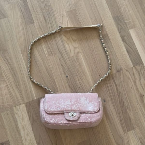 Rosa axelväska med paljetter - Söt rosa axelväska med glittriga paljetter och kedjerem i silver och beige. Väskan har ett klassiskt lock med vridknäppe och är perfekt för dig som vill sticka ut lite extra. 