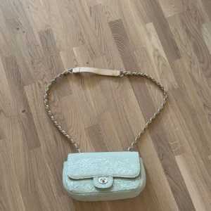 Glittrig beige axelväska med kedja - Snygg beige axelväska med glittriga paljetter och vridbart lås framtill. Väskan har en flätad axelrem i kedja och läderdetalj.