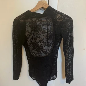 Svart spetsig långärmad bodysuit - Säljer en svart långärmad bodysuit i genomskinlig spets med blommigt mönster. Tight passform och högt skuren nedtill. Perfekt för att skapa en edgy och snygg look. Ingen synlig dragkedja eller knappar, bara ren spets över hela plagget.