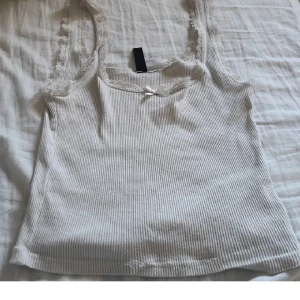 Vit ribbad linnetopp med spets - Supersöt vit ribbad linnetopp med spetskant runt ringningen och axelbanden. Toppen har en liten rosett framtill och är croppad i modellen. Perfekt för varma dagar eller som lager-på-lager-plagg. Materialet är mjukt och stretchigt för en bekväm passform.