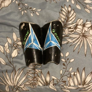 Svarta benskydd från SOC med blått mönster - Säljer ett par svarta benskydd från SOC med blått och vitt geometriskt mönster på framsidan. De har en böjd form för att sitta skönt på benet och insidan är vadderad med svart material för extra skydd. Perfekta för fotbollsträning eller match.