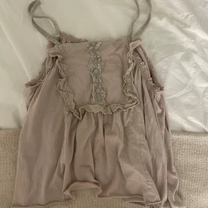 Superfint linne - Supersöt beige linnetopp med smala axelband, volangdetaljer och dekorativa knappar framtill. Toppen har romantiska spetsinslag och en lös, flowy passform som ger en avslappnad vibe. Perfekt att styla med jeans eller kjol för en chill look.