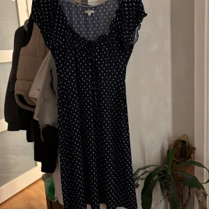 Mörkblå prickig kortklänning - Supersöt mörkblå kortklänning från H&M med vita prickar och volangdetaljer vid ärmarna. Klänningen har en lätt och luftig känsla, perfekt för varma dagar. Snyggt rynkad vid halsen och med korta ärmar för en avslappnad vibe.