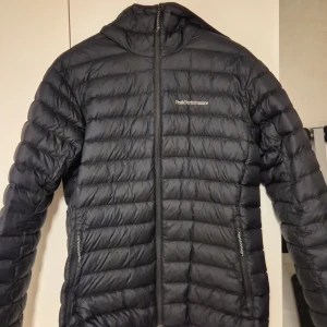 Svart dunjacka Peak Performance - Säljer en svart dunjacka från Peak Performance med huva och dragkedja framtill. Jackan är quiltad med horisontella sömmar och har två fickor med dragkedja. Perfekt för kyliga dagar och enkel att matcha med det mesta.