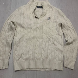 Polo Ralph Lauren tröja  - Kabelstickad vit tröja från Polo Ralph Lauren med halv dragkedja och hög krage. Klassisk design med ribbade muddar och det ikoniska broderade logotypen på bröstet. Perfekt för lager-på-lager och snygg till jeans eller chinos.