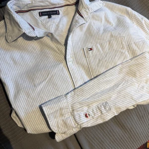 Randig skjorta från Tommy Hilfiger - Snygg skjorta från Tommy Hilfiger i ljusblått och vitt med tunna ränder. Klassisk krage, långärmad och med broderad logga på bröstfickan. Skjortan är i mjuk bomull och har knappar framtill samt vid ärmslut. Perfekt för en clean och stilren look.