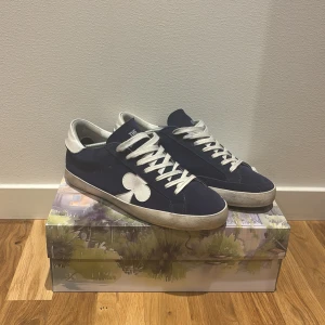 Blå sneakers The Gallerist mocka - Snygga blå sneakers från The Gallerist med vit klöverdetalj på sidan. Skorna har vita snören, vit häl och sula samt ovandel i mocka. Klassisk låg modell med rund tå och platt sula.