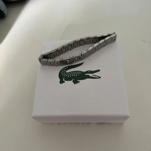 Lacost armband - Stilrent armband från Lacoste i silverfärgad metall med diskreta mönster på länkarna. Kommer i originalask och med tygpåse, perfekt för dig som gillar klassisk och sportig design. Snygg detalj med Lacostes ikoniska krokodillogga.