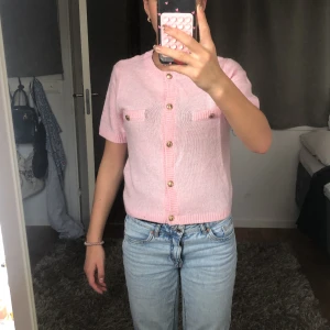 Rosa stickad topp från Gina Tricot - Supersöt ljusrosa stickad topp från Gina Tricot med korta ärmar och snygga guldfärgade knappar framtill. Toppen har två fejkfickor på bröstet och ribbade kanter vid hals, ärmslut och nederkant. Perfekt för dig som gillar en söt och trendig look!