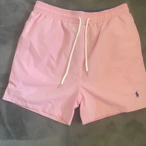 Rosa badshorts från Polo Ralph Lauren - Snygga ljusrosa badshorts från Polo Ralph Lauren med vit snörning i midjan och klassisk blå logga broderad på benet. Shortsen har elastisk midja och innerbyxa i mesh för extra komfort. Perfekta för stranden eller poolen!