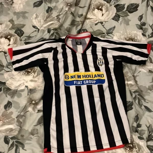 Juventus randig fotbollströja L - Juventus fotbollströja i svart och vitt med klassiska vertikala ränder, korta ärmar och röd kant vid hals och ärmslut. Broderat Juventus-emblem på bröstet och New Holland Fiat Group sponsortryck. Tillverkad i lätt polyester som andas.