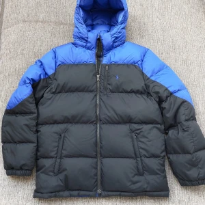 Dunjacka Polo Ralph Lauren - junior - Dunjacka från Polo Ralph Lauren i svart och blått. Jackan har huva, två dragkedjefickor och broderad logga på bröstet. Puffig modell med hel dragkedja framtill. Sparsamt använd och i mycket fint skick! Strlk 170/XL junior