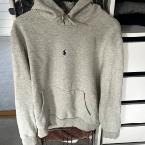 Grå hoodie från Ralph Lauren - Snygg ljusgrå hoodie från Ralph Lauren med klassisk broderad logga på bröstet. Tröjan har huva med snörning, magficka och ribbade muddar. Perfekt för en chill och stilren look.