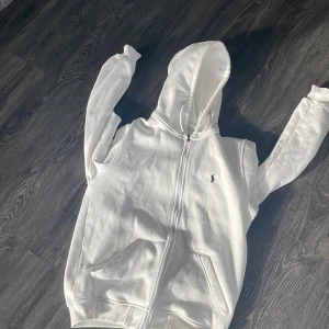 Vit hoodie från Ralph Lauren - Vit hoodie från Ralph Lauren med dragkedja och klassisk blå broderad logga på bröstet. Tröjan har huva, två fickor framtill och är tillverkad i mjuk bomull. Perfekt för en clean och stilren look. Är L men passar xl också 