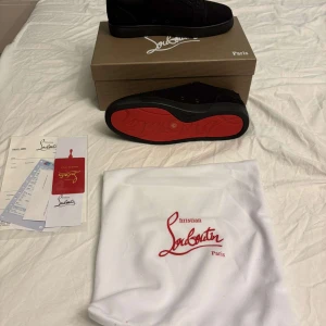 Christian Louboutin svarta sneakers - Säljer ett par svarta sneakers från Christian Louboutin med ikonisk röd sula. Skorna har snörning, rund tå och är tillverkade i mocka med broderad logga på hälen. Kommer med originalkartong, dustbag och äkthetsbevis. Perfekta för dig som vill sticka ut med lyxig streetstyle.