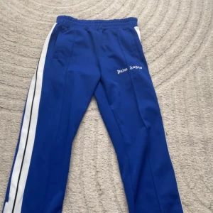 Blå Palm Angels trackpants M - Snygga blå trackpants från Palm Angels med vita sidoränder och logga på låret. Byxorna har elastisk midja och är tillverkade i 100% polyester. Perfekta för en sportig och trendig streetwear-look.