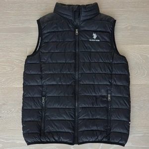 Mörkblå väst från U.S. Polo Assn. - Snygg mörkblå dunväst från U.S. Polo Assn. med broderad logga på bröstet och två dragkedjefickor framtill. Västen är quiltad och har hög krage samt hel dragkedja. Perfekt att slänga över en hoodie för en chill look. Bara att höra av sig vid frågor. 