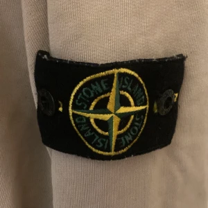 Beige hoodie från Stone Island Junior - Säljer en beige hoodie från Stone Island Junior med klassisk patch på ärmen. Tröjan har huva, magficka och långa ärmar. Perfekt för dig som gillar streetwear och vill ha en stilren look med exklusiv känsla.