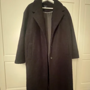 Svart lång teddyjacka från New Look - Mysig svart teddyjacka från New Look med lång passform och klassisk krage. Jackan har två fickor framtill och stängs med knappar. Perfekt för kyliga dagar när du vill ha en stilren och trendig look. Passar S för en oversized look. 