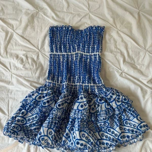 Vit och blå axelbandslös kortklänning - Superfin axelbandslös kortklänning i vitt och blått med broderade detaljer och volangig kjol. Klänningen har smockad överdel och markerad midja, samt ett unikt mönster med blå cirklar och linjer. Perfekt för dig som vill sticka ut med en lekfull och somrig look.