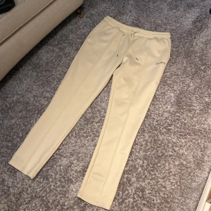 Beige mjukisbyxor från Les Deux - Snygga beige mjukisbyxor från Les Deux med dragsko i midjan och diskret logga på ena benet. Byxorna har en relaxed passform och är tillverkade i mjukt material som känns skönt mot huden. Perfekta för en chill och stilren look. På bilden syns en fläck och därför är dom så billiga.