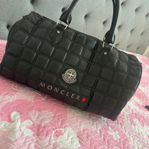 Svart quiltad Moncler handväska - Säljer en svart quiltad handväska från Moncler med rejäla handtag och silverfärgade metalldetaljer. Väskan har ett rutigt mönster i materialet och dragkedja upptill. Perfekt för dig som vill ha en snygg och rymlig väska med exklusiv känsla.